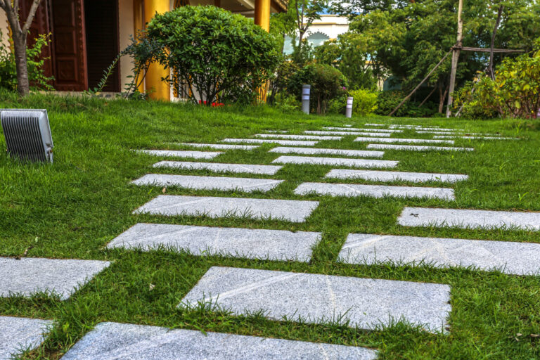 A ground-level, slightly angled view of a garden path made of rectangular gray stone pavers set into a lush green lawn. The path curves gently toward a building with golden columns and a dark wooden door in the background. Mature green bushes and trees surround the lawn, and a small outdoor floodlight is visible in the foreground on the left. The scene is bright and well-manicured, conveying a peaceful outdoor atmosphere.