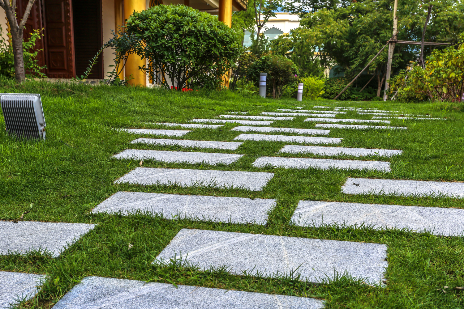 A ground-level, slightly angled view of a garden path made of rectangular gray stone pavers set into a lush green lawn. The path curves gently toward a building with golden columns and a dark wooden door in the background. Mature green bushes and trees surround the lawn, and a small outdoor floodlight is visible in the foreground on the left. The scene is bright and well-manicured, conveying a peaceful outdoor atmosphere.