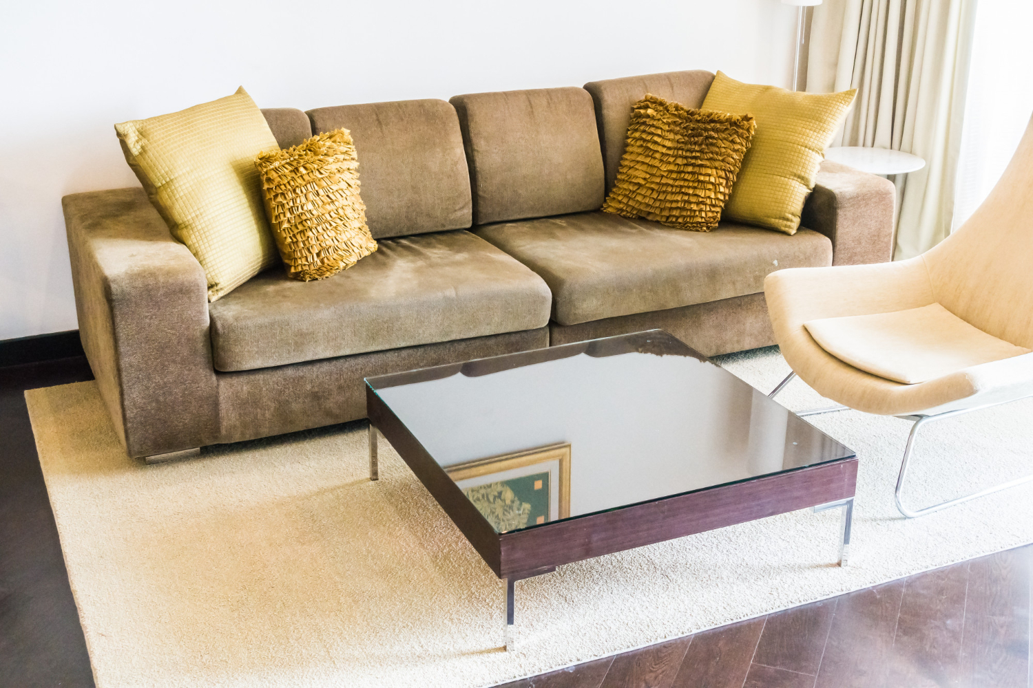 A contemporary living area featuring a plush, golden-brown fabric sofa adorned with four mustard-yellow throw pillows—two smooth and two textured with ruffles. In front of the sofa is a square, dark-framed coffee table with a reflective glass top. To the right, a cream-colored modern lounge chair sits on a light beige rug over dark hardwood flooring.