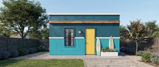 A modern, teal-painted prefabricated house with a bright yellow front door sits in a neatly landscaped backyard. Surfboards lean against the exterior wall next to a white planter box filled with succulents and greenery.